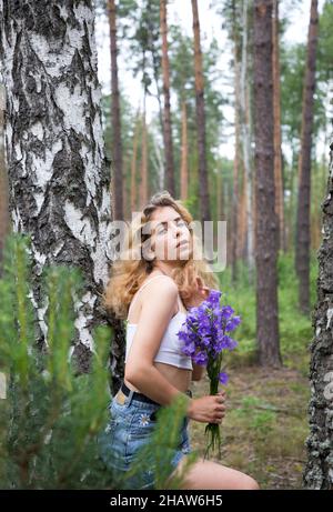 belle adolescente se dresse en forêt parmi les arbres avec un bouquet de fleurs de campanula violet. promenade romantique d'été en forêt, énergie de la nature, rela Banque D'Images