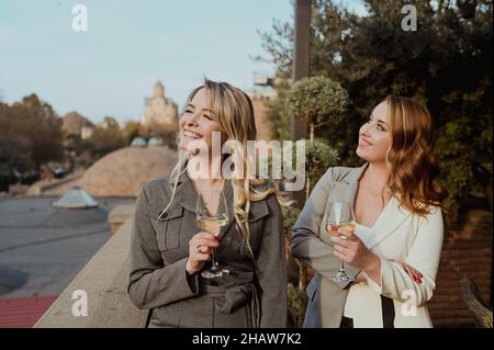 Portrait en gros plan de deux amies en costume strict en train de rire en buvant du vin sur la terrasse à l'extérieur du café de rue d'été sur les bâtiments arrière-plan de la vieille ville de Tbilissi, Géorgie.Photo de haute qualité Banque D'Images