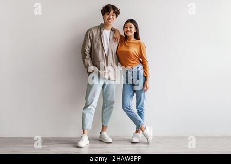Portrait en longueur d'un jeune couple asiatique heureux dans des vêtements décontractés qui s'embrasent et regardent l'appareil photo contre un mur blanc Banque D'Images