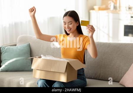Jeune femme asiatique ouvrant la boîte, faisant geste YES et tenant la carte de crédit, satisfait avec l'achat en ligne réussi Banque D'Images