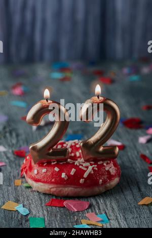 gros plan de deux bougies allumées en forme de nombre doré formant le numéro 22 en ajoutant un petit gâteau avec une glaçage rouge, sur une table rustique grise saupoudrée de bois Banque D'Images