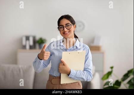 Une psychologue féminine positive se fait le geste du pouce, travaille au bureau et sourit, recommandant un cours de formation Banque D'Images