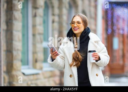 Jeune femme élégante et souriante avec un smartphone et un café en plein air en descendant la rue. Banque D'Images