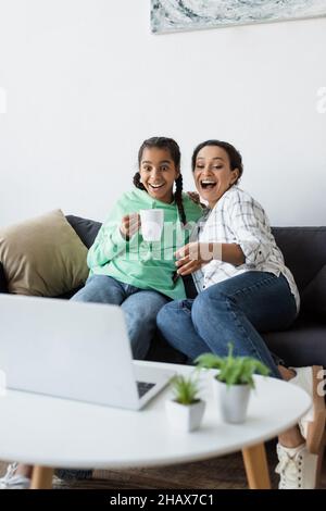 une femme afro-américaine enthousiaste riant et pointant sur un ordinateur portable tout en regardant un film près d'une fille adolescente Banque D'Images