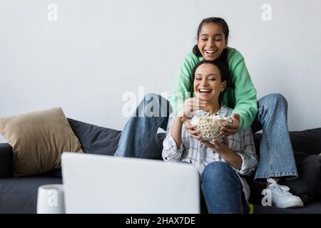 bonne femme afro-américaine avec une fille adolescente regardant un film sur un ordinateur portable près d'un bol de pop-corn Banque D'Images