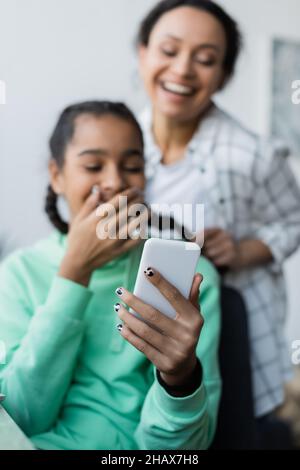 bonne femme afro-américaine qui braque les cheveux de sa fille adolescente en riant tout en regardant son smartphone Banque D'Images