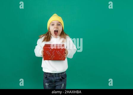 Génial.Un petit enfant excité tient une boîte cadeau et hurle avec joie, bouche ouverte, mur vert Banque D'Images