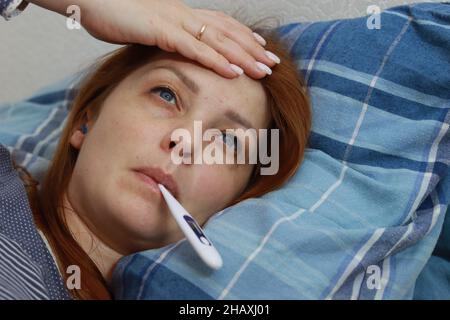 une femme malade est au lit et mesure sa température, un rhume viral et des maux de tête. Banque D'Images