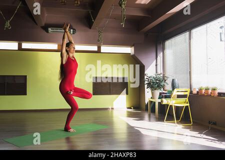 Une femme exécute une variante de vrikshasana exercice, pose d'arbre, se tient sur un tapis dans le studio, s'entraîne dans un rouge sport combinaison Banque D'Images