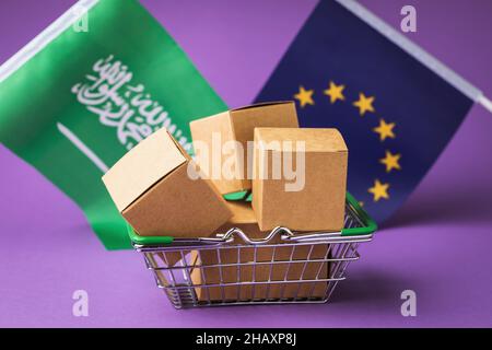 Boîtes en carton dans un panier alimentaire et drapeaux sur fond coloré, le concept de commerce entre l'UE et l'Arabie saoudite Banque D'Images