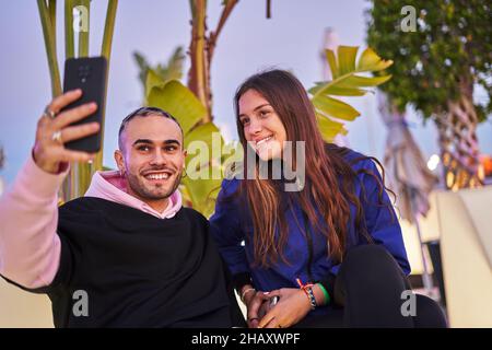 Jeunes amis positifs dans des vêtements décontractés assis près et prenant autoportrait sur le téléphone portable au coucher du soleil Banque D'Images