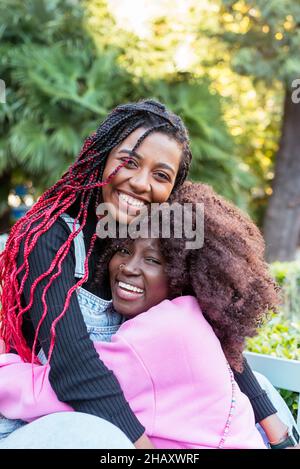 De jeunes amies afro-américaines s'embrassent les unes avec les autres tout en étant assis sur le banc dans le parc et en regardant la caméra avec plaisir Banque D'Images