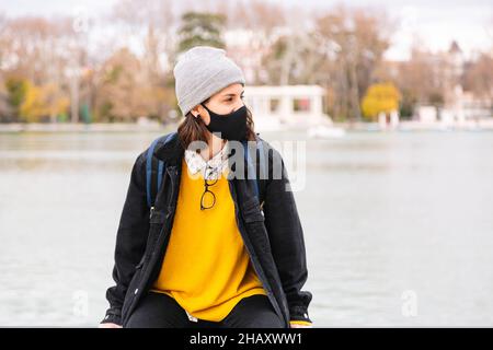 Jeune femme voyageur en vêtements d'extérieur et avec sac à dos et portant un masque de protection du visage assis sur la clôture du lac Banque D'Images