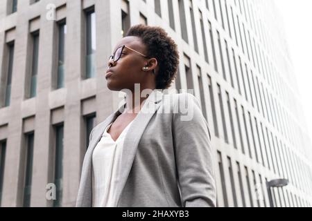Faible angle de jeune femme entrepreneur en veste élégante et lunettes de vue debout près des gratte-ciels modernes de la ville Banque D'Images