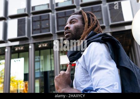 Jeune homme noir barbu avec des dreadlocks portant une chemise blanche tenant la veste au-dessus de l'épaule tout en regardant loin dans un immeuble de bureau moderne Banque D'Images