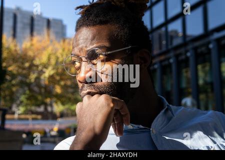 Homme américain d'Afrique sérieux en lunettes et décontracté porter avec des dreadlocks et la main sur le menton près du mur de verre du bâtiment Banque D'Images