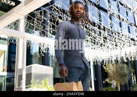 Faible angle d'homme noir confiant avec des dreadlocks et dans des vêtements en denim avec des sacs en papier près du centre commercial avec des guirlandes Banque D'Images