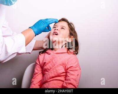 Un médecin non reconnaissable est présent dans les gants en latex. Il prélève un échantillon de salive de la bouche d'un petit patient tout en effectuant un test de coronavirus contre le backgroun gris Banque D'Images