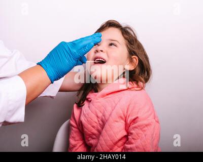 Un médecin non reconnaissable est présent dans les gants en latex. Il prélève un échantillon de salive de la bouche d'un petit patient tout en effectuant un test de coronavirus contre le backgroun gris Banque D'Images