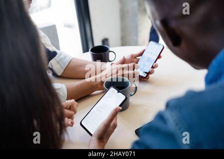 Groupe de collègues multiraciaux non reconnaissables et écourté surfant sur des téléphones portables tout en étant assis à table avec des boissons chaudes pendant une pause dans un café moderne et lumineux Banque D'Images