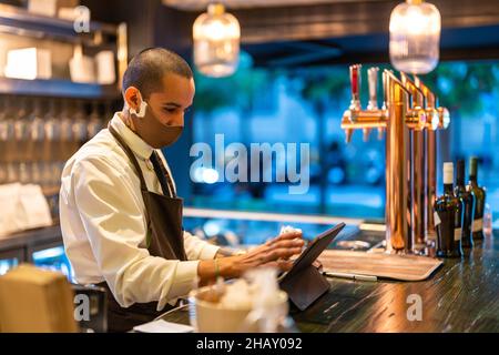 Barkeeper mâle sérieux en uniforme et masque travaillant sur un comprimé à l'opposé de la barre pendant une pandémie de coronavirus Banque D'Images