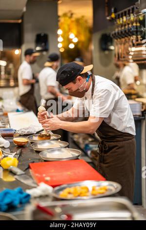 Vue latérale d'un cuisinier professionnel dans un masque de protection en ajoutant de la crème à partir d'un sac de pâtisserie tout en préparant un délicieux hamburger dans la cuisine du restaurant au travail Banque D'Images