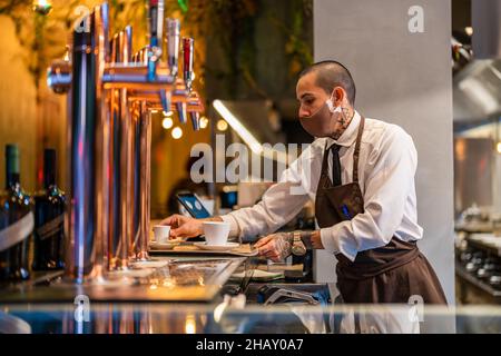 Serveur positif en uniforme et masque plaçant des tasses de café filtre aromatique sur le plateau au comptoir avec le clavier de la station d'accueil dans le restaurant pendant le coronavir Banque D'Images