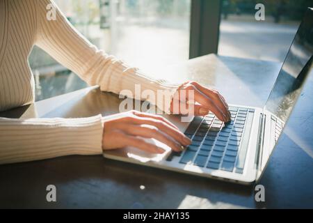 Crochez une femme anonyme dans un chandail chaud assis à table dans un café et parcourant un ordinateur portable tout en travaillant sur le projet à distance dans la lumière du jour Banque D'Images