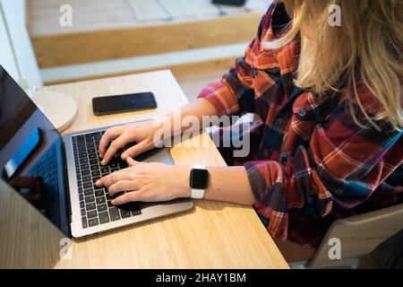 Courte femme non reconnaissable dans une chemise à carreaux assise sur une table en bois et dactylographiant sur un ordinateur portable dans la chambre à la mansarde dans le village le matin ensoleillé Banque D'Images