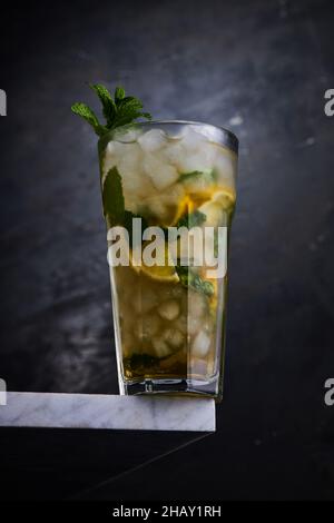 Verre transparent de cocktail d'alcool avec des tranches de citron et de feuilles de menthe verte avec de la glace servi sur le coin de la table sur fond gris Banque D'Images