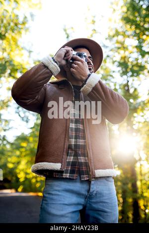 Photo de l'homme ci-dessous en tenue élégante en prenant des photos avec un appareil photo d'époque de la nature tout en se tenant sur un chemin d'asphalte près de grands arbres verts pendant Banque D'Images