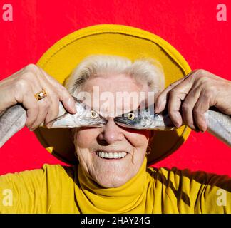 Femme âgée positive avec cheveux gris dans un élégant chapeau jaune couvrant les yeux avec du poisson cru tout en se tenant sur fond rouge Banque D'Images