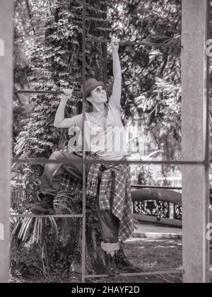 Une dame vêtu d'un chapeau et d'une grande tenue de rue grimpe une clôture en métal dans un parc ombragé.Photo en noir et blanc Banque D'Images