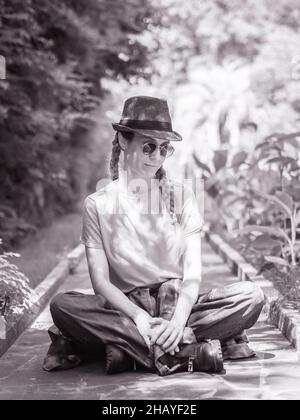 Une dame aux pieds de porc dans des lunettes de soleil et un chapeau est assise à pieds croisés sur le trottoir dans le parc d'été.Photo en noir et blanc Banque D'Images