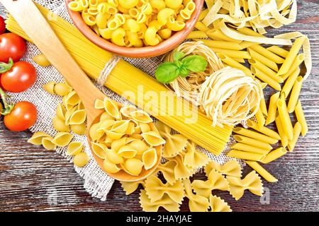 Pâtes de coquillages dans une cuillère, tuyau de rigate dans un bol, spaghetti, tagliatelle, penne, farfalle,transtette, tomates et persil sur toile de jute sur bac en bois Banque D'Images