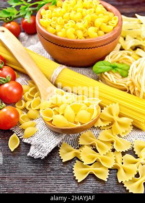 Pâtes de coquillages dans une cuillère, pipes de rigate dans un bol, spaghetti, tagliatelle, farfalle, trenette,tomates et persil sur toile de fond en bois Banque D'Images