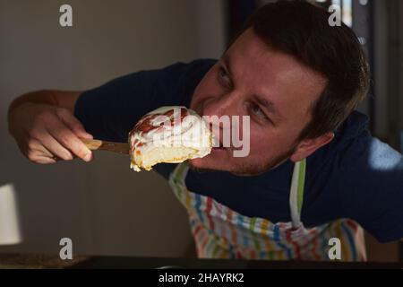 Un homme dans un tablier goûte des cinnamons faits maison dans la cuisine.Vue avant. Banque D'Images