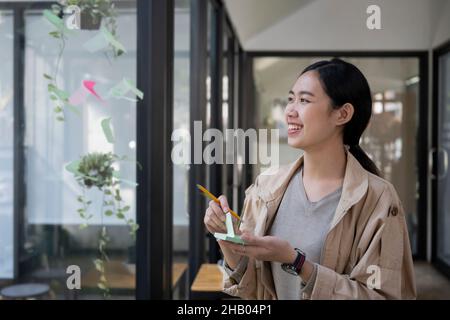 Femme créative souriante lisant des notes collantes sur un mur de verre dans un lieu de travail moderne. Banque D'Images