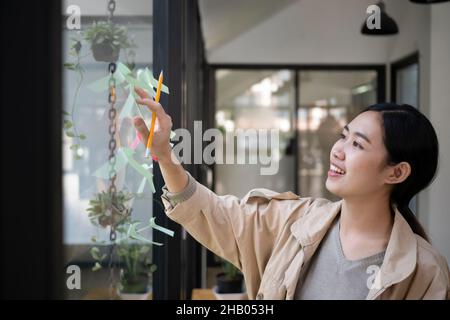 Jeune femme asiatique souriante lisant des notes adhésives sur un mur de verre dans un bureau créatif. Banque D'Images