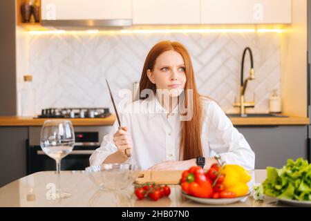 Portrait d'une jeune femme à tête rouge attrayante avec des yeux insatisfaits tenant un gros couteau assis à la table avec planche à découper, en regardant loin Banque D'Images