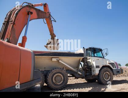 pelle hydraulique à chenilles orange et tombereau articulé pendant l'excavation dans une fosse de sable par une chaude journée ensoleillée. chargement et transport du sol Banque D'Images