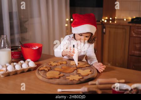 Petite fille à chapeau santas décorent le pain d'épice à la maison.Concept de traditions de Noël et du nouvel an.Boulangerie de Noël.Joyeuses fêtes Banque D'Images