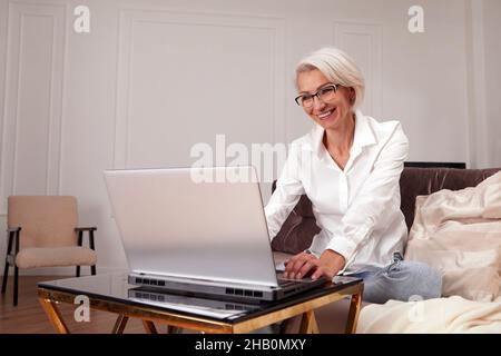 Bonne 50 plus âgée adulte d'âge moyen, femme tenant un ordinateur portable assis sur un canapé à la maison.Femme élégante et souriante aux cheveux gris en verre Banque D'Images