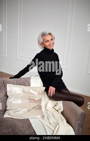 Angle élevé de la femme d'âge moyen positive avec cheveux gris et dans une tenue noire tendance assis sur un canapé dans la chambre et regardant l'appareil photo Banque D'Images