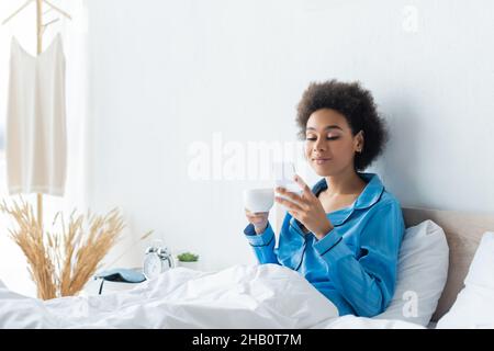 femme afro-américaine souriante en pyjama utilisant un téléphone portable et tenant une tasse de café dans le lit Banque D'Images