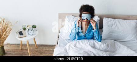 vue en grand angle de la femme afro-américaine qui ajuste le masque de sommeil dans la chambre, la bannière Banque D'Images