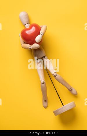 Une petite figurine en bois épouse délicatement une tomate rouge sous la forme d'un cœur, symbole de l'amour, sur fond jaune.Vue de dessus. Banque D'Images