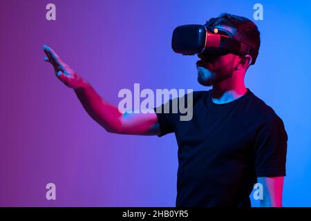 Portrait d'un homme utilisant un casque VR tout en communiquant avec un ami dans un métaverse dans un intérieur sombre lumière néon éclairée.Gros plan futuriste Banque D'Images