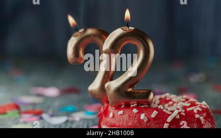 bougies illuminées en forme de nombre doré formant le numéro 22 sur un petit gâteau avec une glaçage rouge, sur une table en bois rustique gris avec un peu de confetti, dans une casserole Banque D'Images