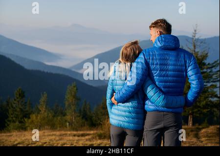 Vue arrière des amoureux qui s'embrasent et se regardent les uns les autres sur fond de magnifiques enfants de montagne, tout en marchant dehors dans le temps ensoleillé du début de l'automne. Banque D'Images